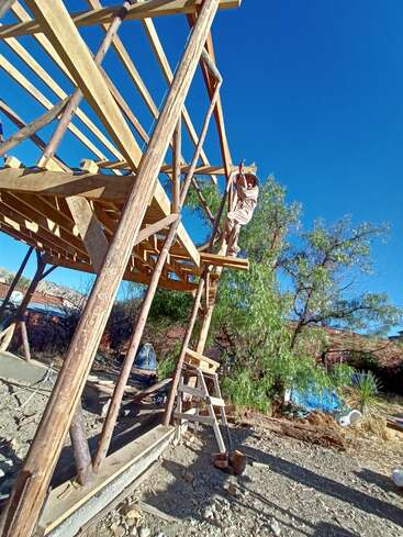 Eine Person steht auf einer im Bau befindlichen Holzkonstruktion und arbeitet mit Werkzeugen unter einem klaren blauen Himmel, umgeben von Bäumen, Felsen und verstreuten Baumaterialien.