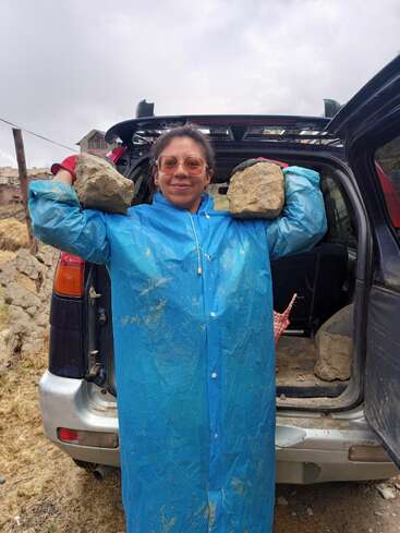 Eine Frau in einem blauen Regenmantel und mit einer rosafarbenen Brille hält zwei große Steine auf den Schultern. Sie steht an einem bewölkten Tag im Freien neben einem offenen SUV-Kofferraum.