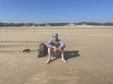 A person sits on a sandy beach under a bright blue sky, wearing a cap and colorful shirt, with a bag, bottle, and shoes nearby.