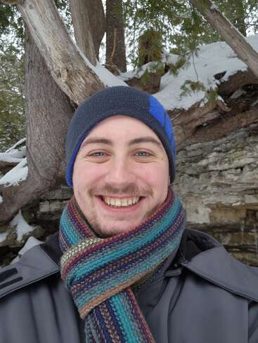 A smiling person in winter attire, wearing a striped scarf and beanie, stands outdoors near snow-covered trees and rocks, enjoying a cold, scenic environment.