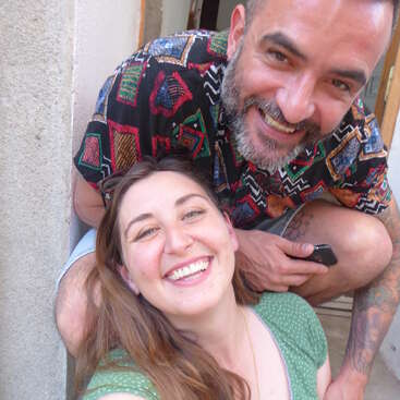 A man and woman smile brightly at the camera. The man has a beard and patterned shirt; the woman wears green and sunglasses, both appear very happy.