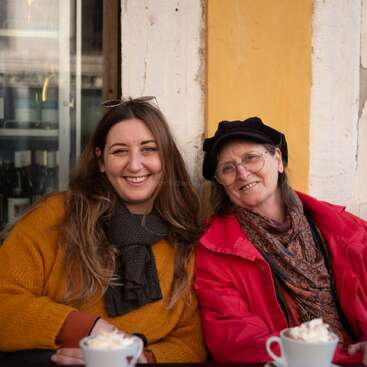 Two women sit closely together at a café, smiling warmly. They wear cozy scarves and jackets. Hot beverages topped with whipped cream are on the table before them.