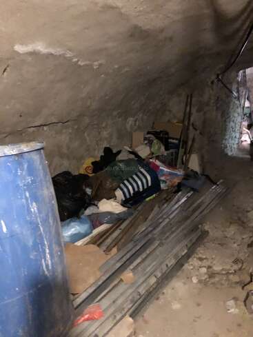 L'image montre un tunnel souterrain encombré et faiblement éclairé, avec des murs en pierre, des tiges métalliques, un grand tonneau bleu, divers sacs, vêtements, boîtes et débris épars.