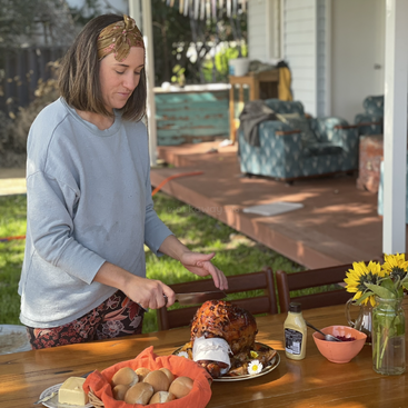 A woman stands at a wooden table, carving a glazed ham with a knife, surrounded by a basket of rolls, flowers, and other food items on a sunny day.