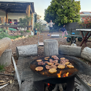 The image depicts a backyard scene with a fire pit containing meat, surrounded by logs and a covered patio area, with people and children in the background.