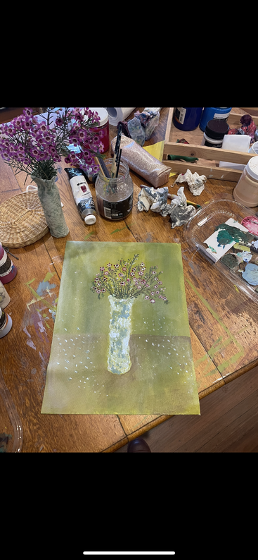 The image depicts a person\'s hands holding a paintbrush, with a canvas featuring a painting of a vase and flowers in the background, set against a light-colored wall.