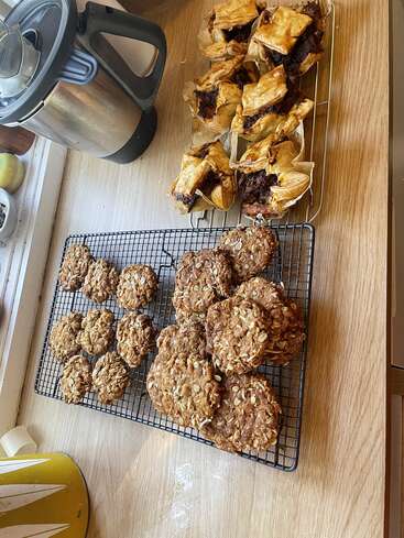 The image depicts a kitchen countertop with a coffee maker, a tray of cookies, and a tray of pastries on a wooden surface.