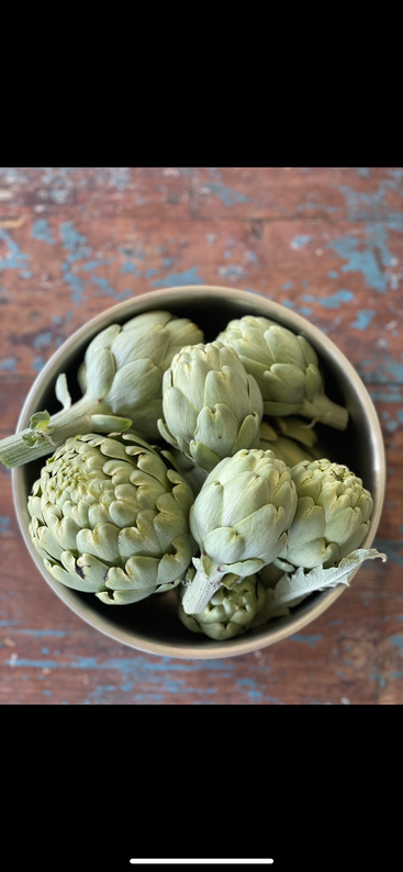 The image depicts a close-up of a pile of artichokes, with their leaves and stems visible, set against a light-colored background. The artichokes are arranged in a neat and organized manner.