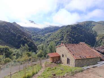 A rustic stone house with a red-tiled roof sits on a hillside, surrounded by lush, green mountains, under a partly cloudy, blue sky.