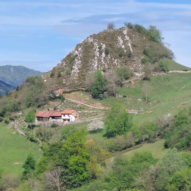 The image depicts a serene rural landscape featuring a small house with a red roof, nestled among lush green hills and trees, under a blue sky with wispy clouds.