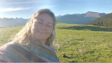 A woman with wavy blonde hair smiles, wearing a colorful poncho. She stands in a sunlit green field with majestic mountains and clear blue sky behind her.