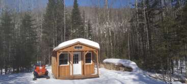 In einem verschneiten Wald steht eine kleine Holzhütte mit schneebedecktem Dach. Daneben stehen ein rotes Schneemobil und ein großer Stapel Brennholz.