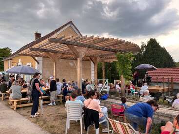 Bei einem lebhaften Treffen im Freien unterhalten sich die Leute unter einer hölzernen Pergola neben einem rustikalen Gebäude, während auf einer kleinen Bühne Live-Musik gespielt wird und der Himmel bewölkt ist.