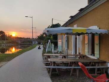 Ein Uferweg bei Sonnenuntergang, neben einem rustikalen Gebäude mit Holzbänken unter einem Vordach. Spiegelungen, sanftes Licht, eine ruhige Szene, eine Person in der Ferne.