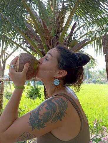 Una mujer con un tatuaje floral y pendientes de flores azules bebe de un coco bajo una palmera, rodeada de exuberante vegetación y un campo de arroz.