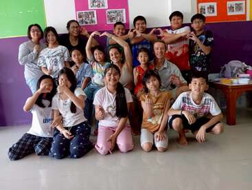 Un alegre grupo de niños y adultos posa en el interior, haciendo formas de corazón con las manos, sonriendo alegremente, lo que sugiere un entorno amistoso y solidario en el aula o en la comunidad.