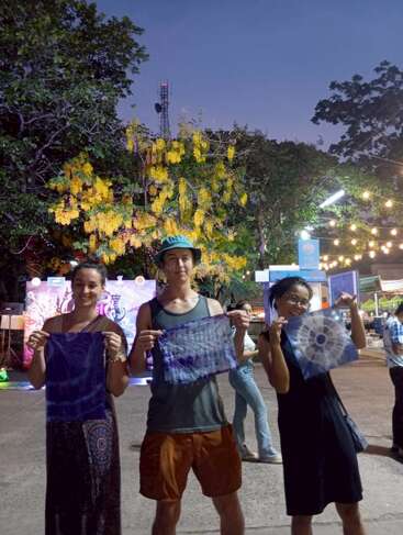 Tres personas están de pie al aire libre al atardecer, sonriendo y sosteniendo con orgullo cuadrados de tela teñida. Las luces decorativas, el árbol de flores amarillas y el vibrante ambiente del mercado crean un animado telón de fondo.