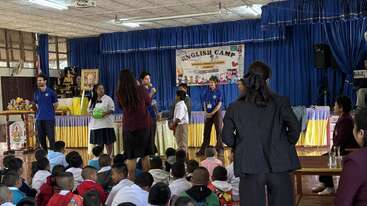 A group of children and adults participate in activities on stage at an English camp event, while an audience of seated children watch attentively in a hall.