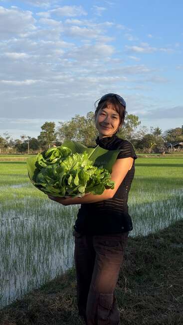 A woman smiles while holding a basket of fresh green lettuce, standing in front of a lush, green rice field under a bright blue sky.
