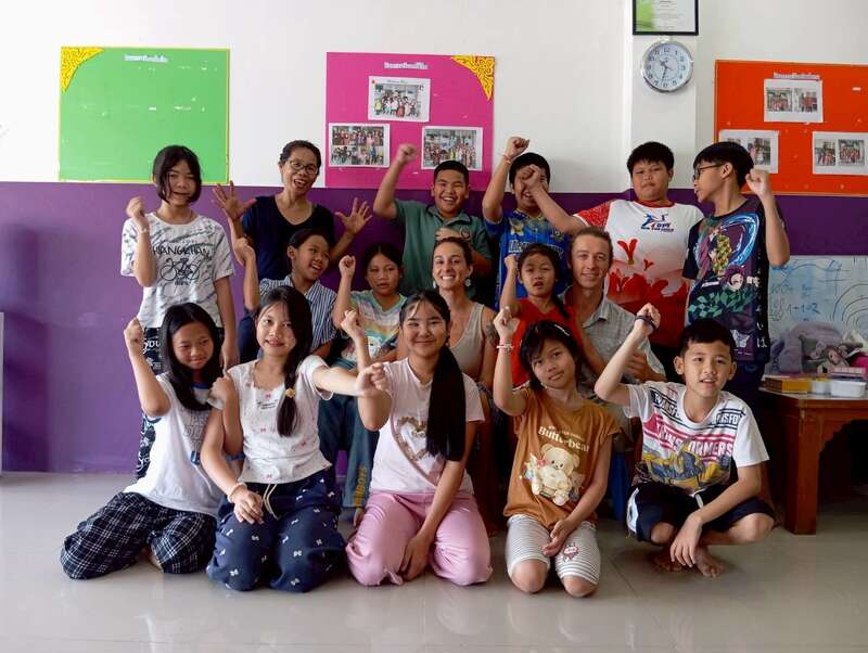 Un grupo de niños y adultos sonrientes posan juntos en una colorida aula. Parecen alegres y enérgicos, haciendo signos de la paz y levantando las manos con entusiasmo.