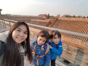Das Bild zeigt eine Frau und zwei Kinder, die für ein Selfie auf einer Brücke mit Geländer posieren, vor dem Hintergrund traditioneller chinesischer Architektur.