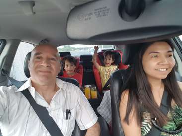 Das Bild zeigt eine vierköpfige Familie in einem Auto, mit zwei Kindern auf dem Rücksitz und zwei Erwachsenen vorne, die alle lächeln und in die Kamera schauen.