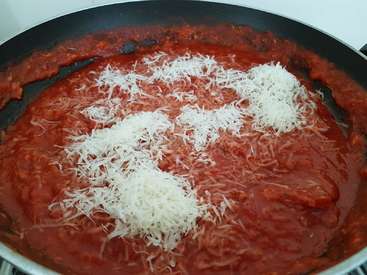 Das Bild zeigt eine große Pfanne mit einer Tomatensoße und geriebenem Käse vor einem weißen Hintergrund.