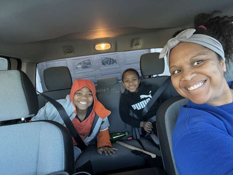 Das Bild zeigt eine Frau und zwei Kinder auf dem Rücksitz eines Autos, wobei die Frau rechts und die Kinder in der Mitte sitzen.