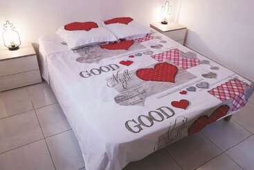 Un lit soigneusement fait avec un drap blanc décoré de cœurs rouges et gris, le texte « Good Night », deux oreillers et des tables de chevet minimalistes avec des lampes modernes.