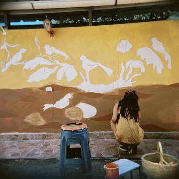 A woman in a yellow dress paints a mural of birds and plants on a wall. Art supplies and a blue stool are scattered nearby.