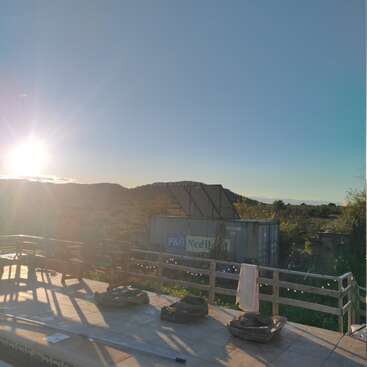 The photo shows a sunlit outdoor scene with hills, a shipping container, wooden railings, a towel, construction materials, and a clear blue sky.