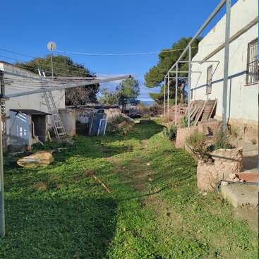 A backyard area with green grass, old structures, metal frames, and various construction materials scattered around, under a bright blue sky with some trees.