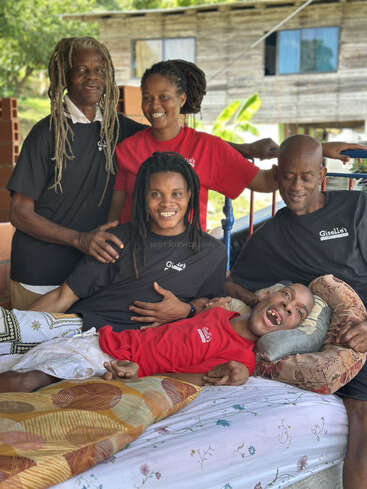 Five people are gathered together, smiling warmly. One person lies on a bed, supported by the others. The background shows a rustic house and greenery.