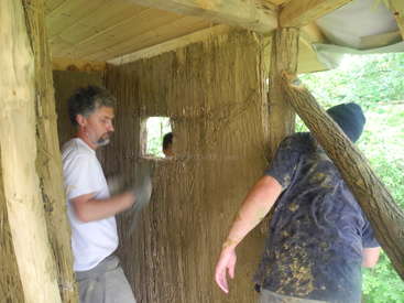 La imagen muestra a dos hombres construyendo una estructura de madera, posiblemente una casa en un árbol, uno de ellos con camisa blanca y el otro con una camisa azul oscuro cubierta de barro.