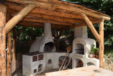 La imagen representa un gran horno de exterior con techo metálico, rodeado por un muro de piedra, con puerta de madera y tirador metálico, enmarcado en un paisaje natural.