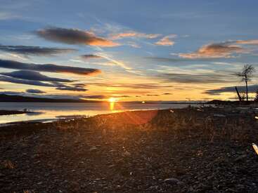 A imagem mostra um pôr do sol tranquilo à beira do lago, com a luz dourada do sol refletindo nas águas calmas, nuvens espetaculares e uma costa rochosa, criando uma atmosfera pacífica e pitoresca.