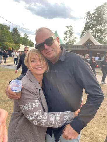 Uma mulher sorridente abraça um homem de óculos escuros em um evento ao ar livre. Ela segura uma bebida, e pessoas e tendas são visíveis no animado fundo.