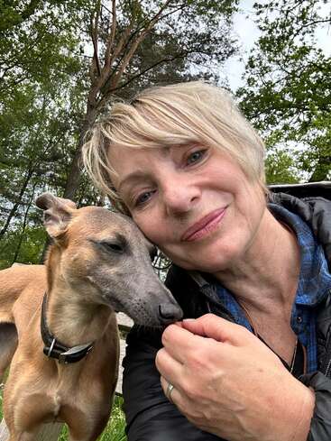 Eine Frau mit kurzen blonden Haaren macht draußen ein Selfie aus nächster Nähe, während sie ihren hellbraunen Hund im Arm hält und sanft lächelt. Bäume und Grünflächen bilden den friedlichen Hintergrund.