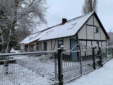 Hinter einem schwarzen Metallzaun, umgeben von Bäumen, steht ein charmantes, traditionelles Haus mit steilem Dach. Überall bedeckt Schnee das Dach, den Boden und die kahlen Äste.