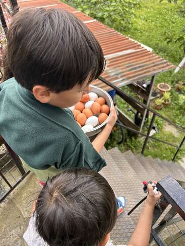 Zwei Kinder kommen eine Metalltreppe im Freien herunter. Das ältere Kind hält vorsichtig eine Schale mit braunen und weißen Eiern in der Hand; das jüngere Kind hält ein Spielzeug in der Hand, möglicherweise eine Wasserpistole.