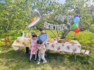 Drei Kinder posieren fröhlich vor einem festlich gedeckten Geburtstagstisch im Freien, auf dem Essen, Luftballons und ein „Happy Birthday“-Schild stehen, umgeben von grünen Bäumen und Sonnenschein.