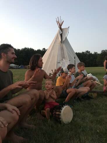 A group of adults and children sit outside near a tipi, clapping and smiling, enjoying music together in a relaxed, natural outdoor setting at dusk.