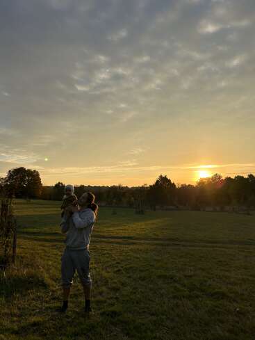 A person holds a baby in an open grassy field at sunset. The sky is partly cloudy with golden hues, creating a peaceful, warm, and serene atmosphere.