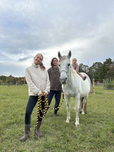 Three people, including a small child sitting on a white horse, are outdoors in a grassy field. They look happy and are enjoying a pleasant day.