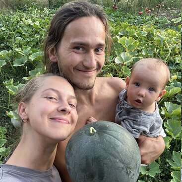 A smiling couple and baby pose in a lush garden, holding a large green melon, surrounded by leafy plants and sunlight, Radiating happiness and togetherness.