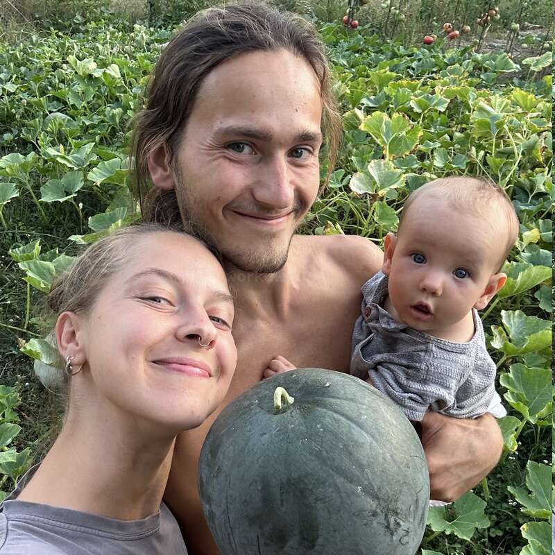 A smiling couple and baby pose in a lush garden, holding a large green melon, surrounded by leafy plants and sunlight, Radiating happiness and togetherness.