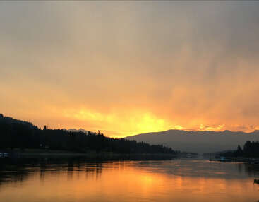 Un coucher de soleil serein projette des teintes dorées sur une rivière calme, entourée de collines sombres couvertes d'arbres et de montagnes lointaines, dans une atmosphère paisible et sereine.