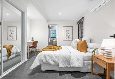 Dieses moderne Schlafzimmer verfügt über ein gemütliches Bett mit weißer Bettwäsche und senfgelben Akzenten, einen hölzernen Nachttisch, einen verspiegelten Kleiderschrank, einen Arbeitsbereich am Schreibtisch und minimalistische Wandkunst.