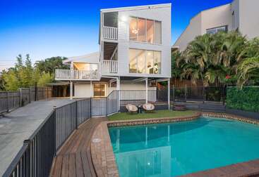 Das moderne dreistöckige Haus mit großen Fenstern und Balkonen hat einen Blick auf einen eingezäunten Pool. Der Garten ist mit einer tropischen Landschaft, Holzterrassen und Sitzgelegenheiten im Freien ausgestattet, die eine entspannte Atmosphäre schaffen.