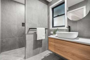 Dieses moderne Badezimmer hat große graue Fliesen, eine begehbare Dusche, einen hölzernen Waschtisch mit Gefäßwaschbecken, einen runden Spiegel, schwarze Armaturen, ein Fenster und ein ordentlich gefaltetes weißes Handtuch.